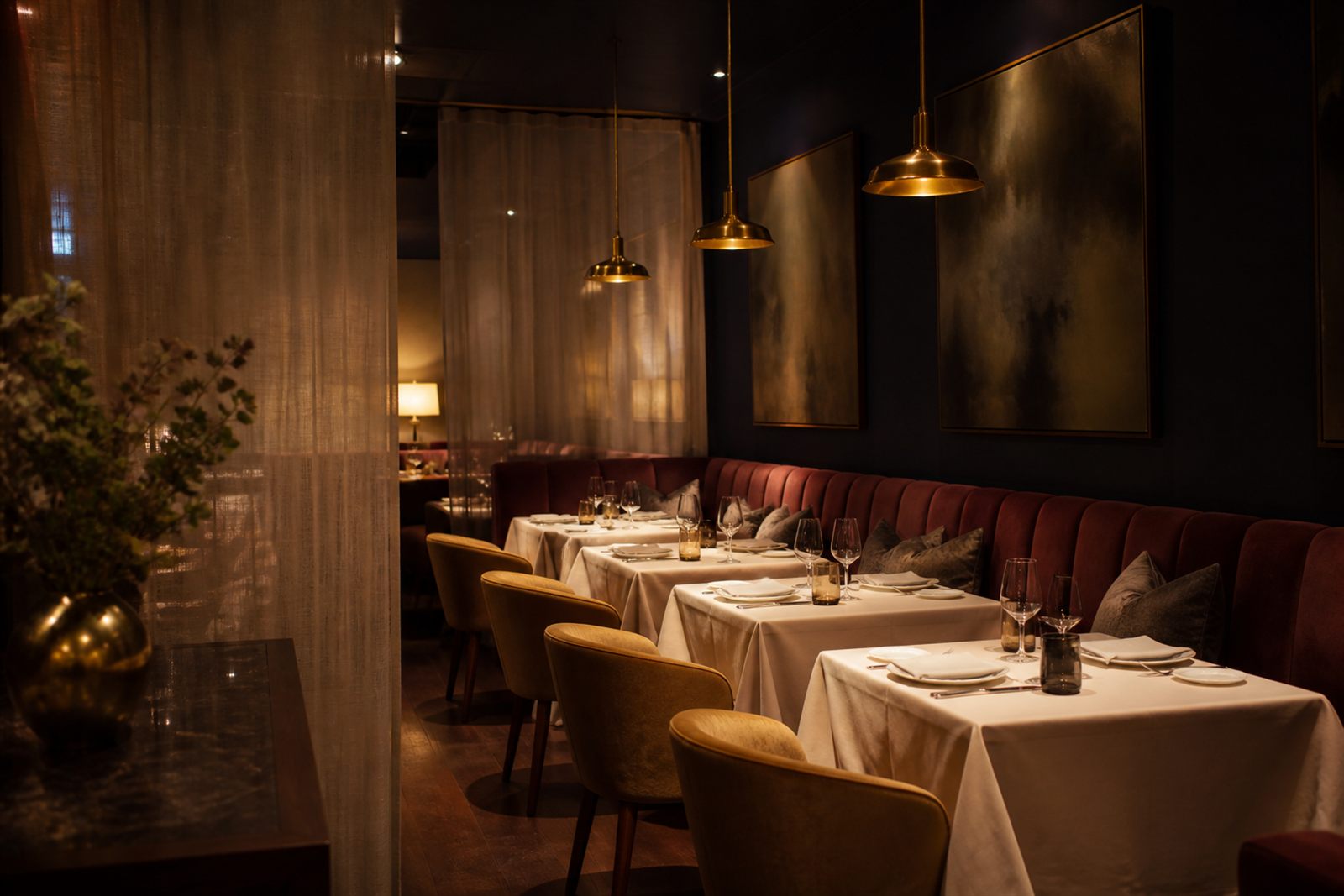 Restaurant dining room with an oxblood velvet banquette against a midnight wall, cream linen-topped tables, gold upholstered chairs, and low brass pendant lights.